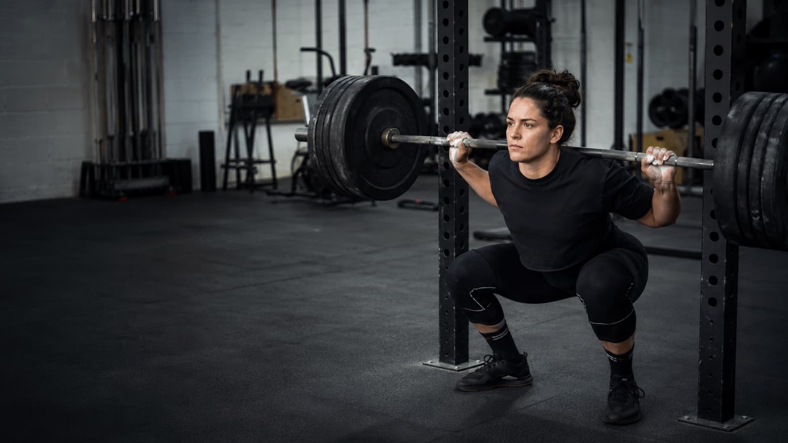 Documentary-style photo of a lifter at the bottom of a heavy barbell back squat in a squat rack, three-quarter front angle, thighs at parallel, in a free-weights gym with the background softly out of focus.